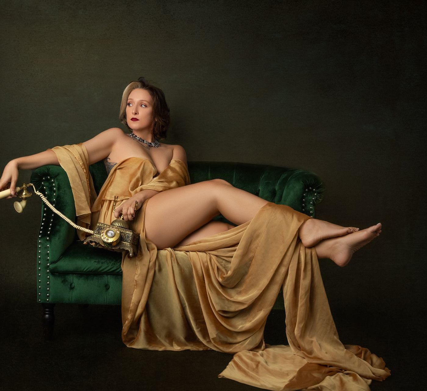 Woman in golden silk fabric poses elegantly on a green velvet couch during a fine art boudoir photoshoot
