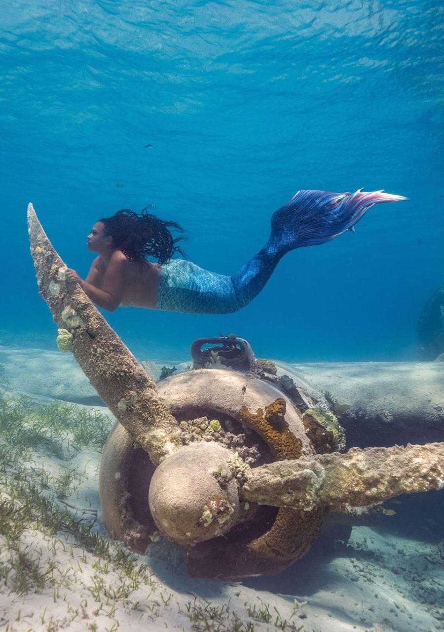 Mermaid with a blue tail posing beside a sunken anchor in shallow salt water