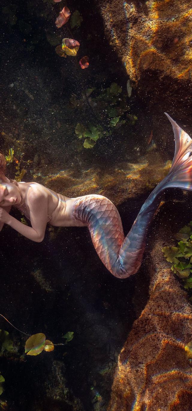 Mermaid in a scaled tail lying among rocks and plants in a spring cavern