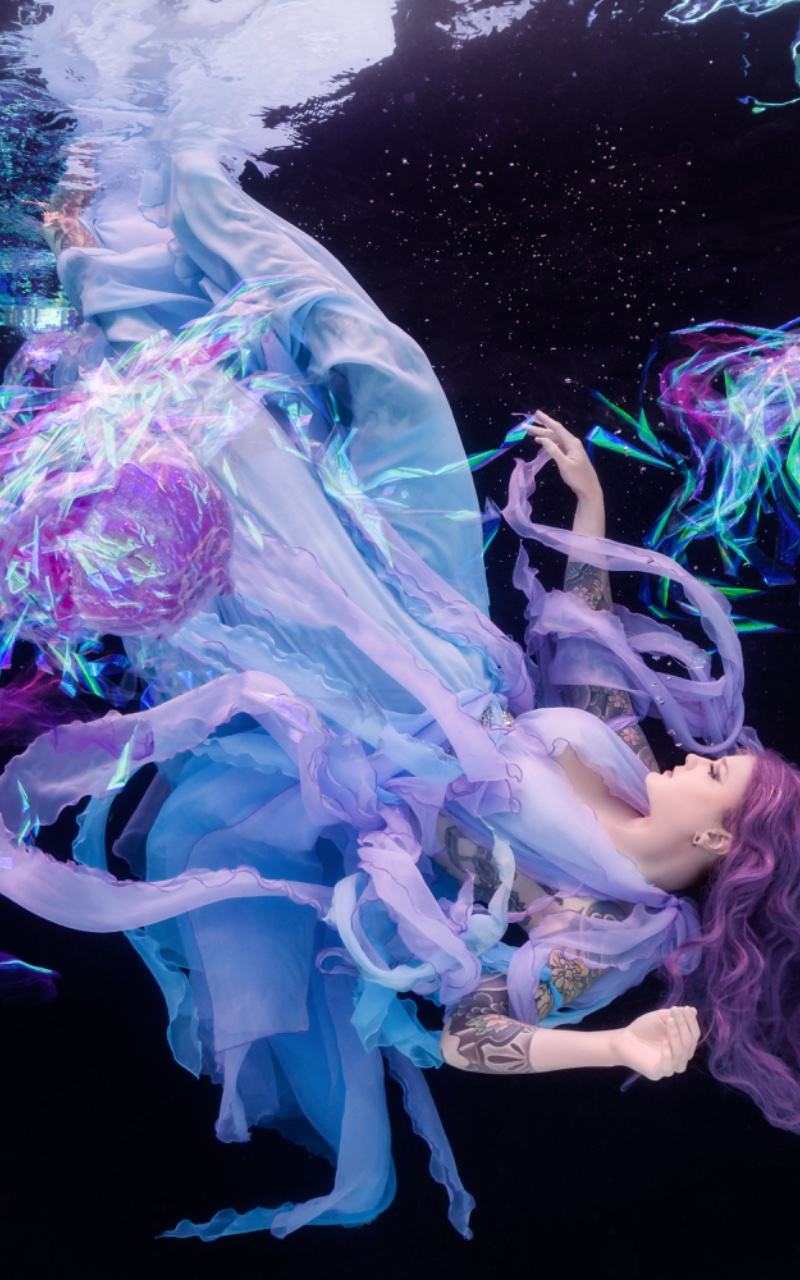 Model in a flowing gown floating underwater surrounded by glowing jellyfish