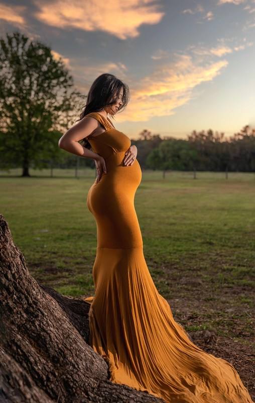 Pregnant woman in golden dress poses outdoors at sunset during a maternity photoshoot