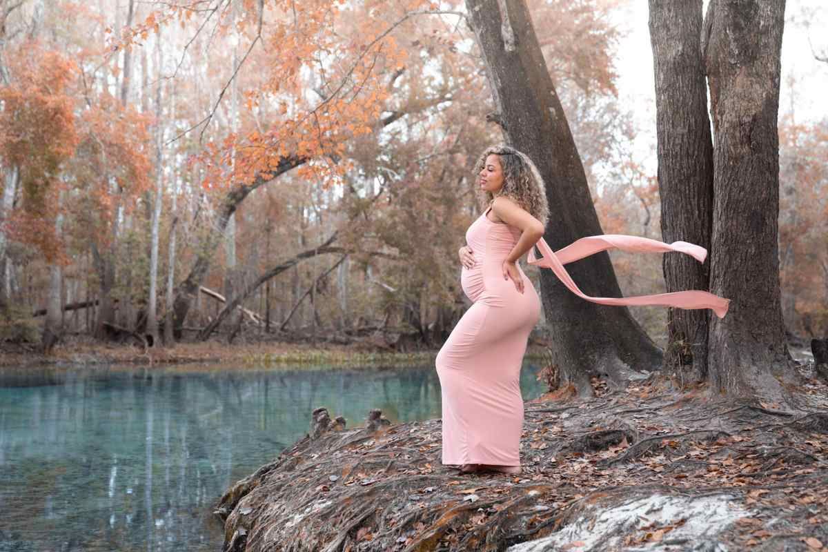 Pregnant woman in pink dress poses outdoors by a turquoise spring during a maternity photoshoot with Water Bear Photography
