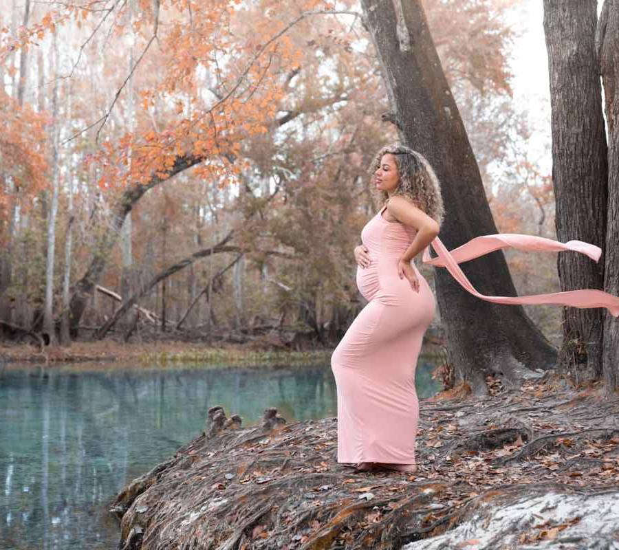 Pregnant woman in pink dress poses outdoors by a turquoise spring during a maternity photoshoot with Water Bear Photography