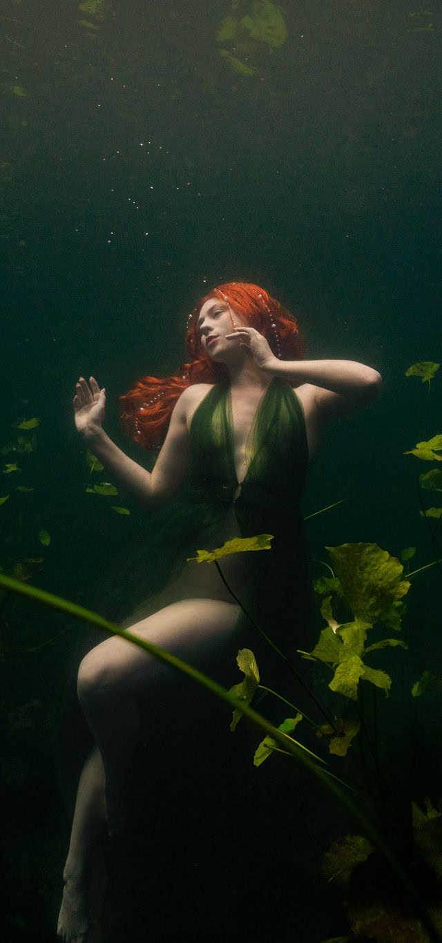 Model with red hair poses among lily pads underwater in a cenote