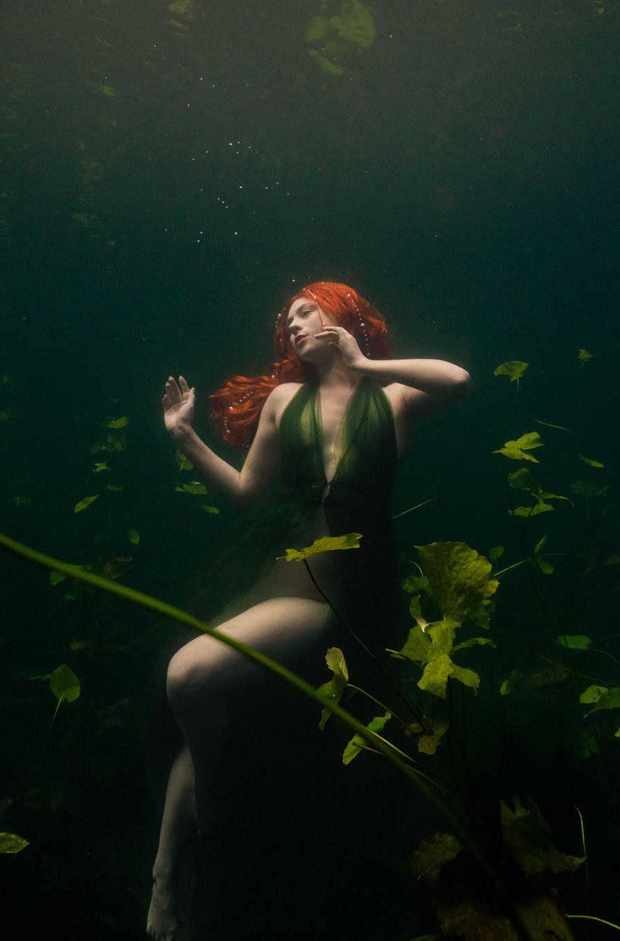 Model with red hair poses among lily pads underwater in a cenote