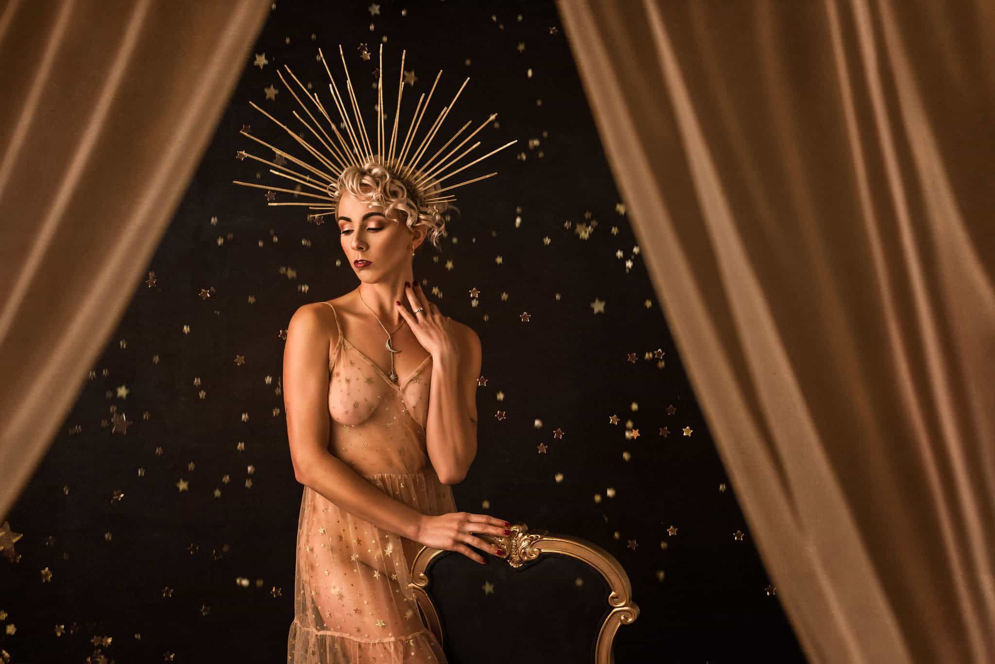 An old Hollywood style studio boudoir photoshoot of a model standing next to a chair wearing a dress covered in gold stars