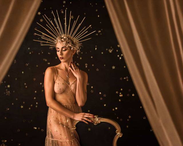 An old Hollywood style studio boudoir photoshoot of a model standing next to a chair wearing a dress covered in gold stars