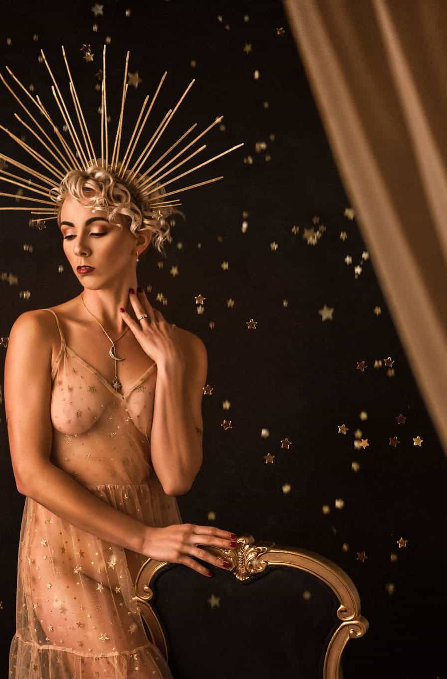 An old Hollywood style studio boudoir photoshoot of a model standing next to a chair wearing a dress covered in gold stars