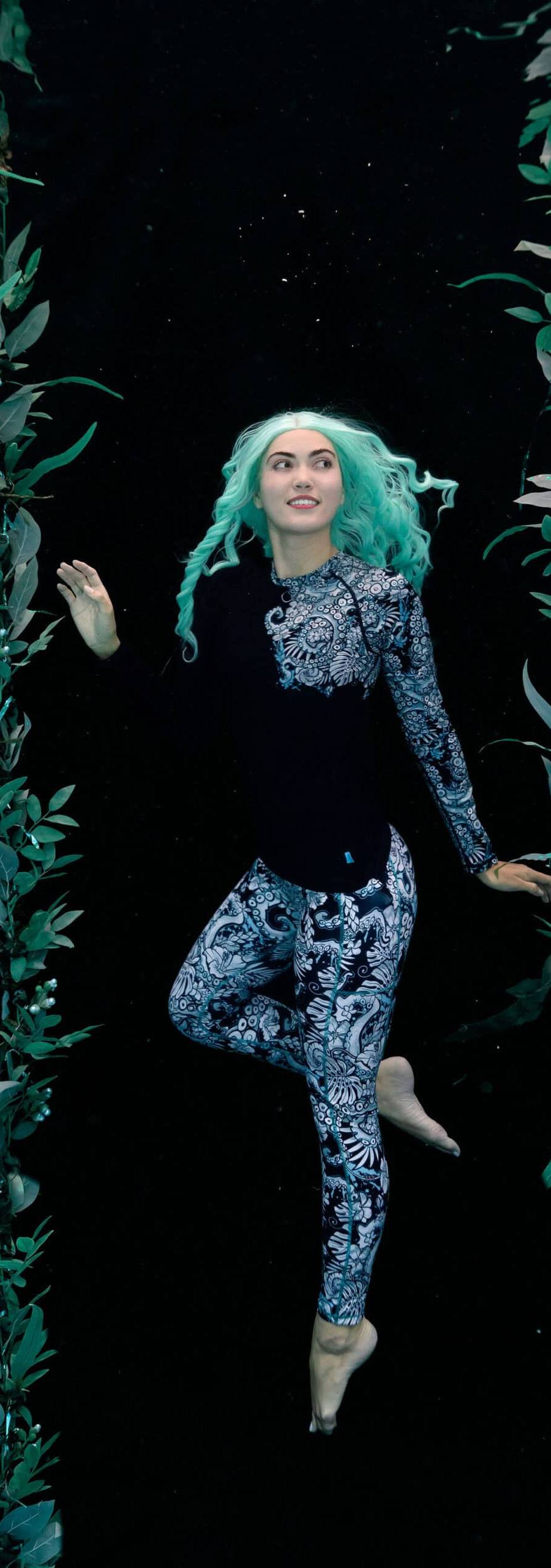 Model poses underwater against a backdrop with kelp around her while wearing black and white rashguard and pants by Spacefish Army.