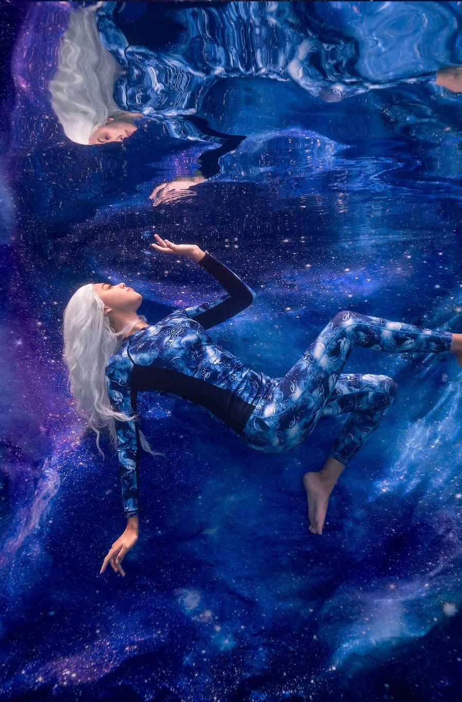 Model posing underwater with a galaxy background as she interacts with her reflection on the water's surface.