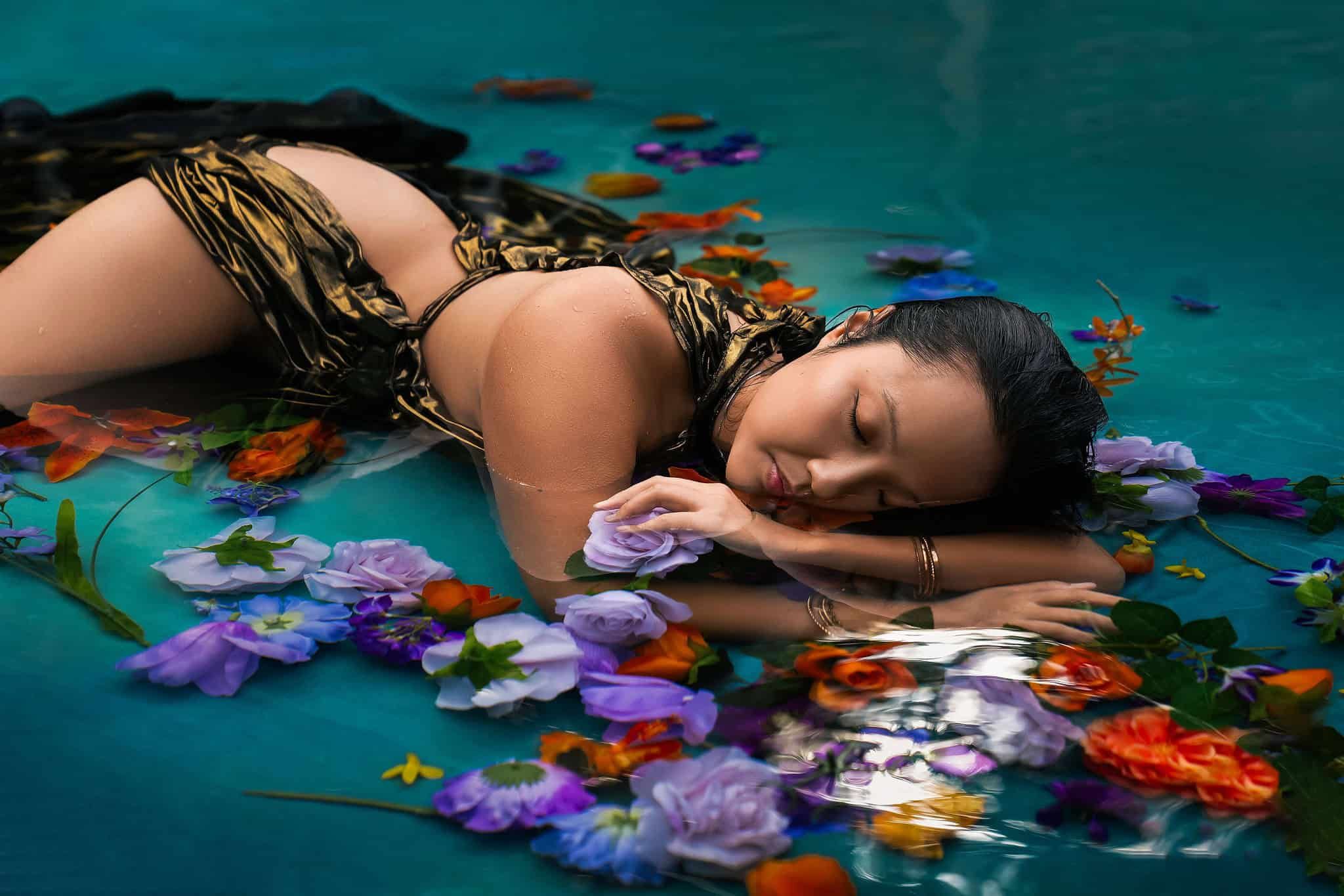 Woman lay on the surface of the water surrounded by flowers wearing a gold dress during a water boudoir photoshoot