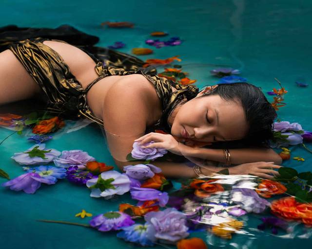 Woman lay on the surface of the water surrounded by flowers wearing a gold dress during a water boudoir photoshoot