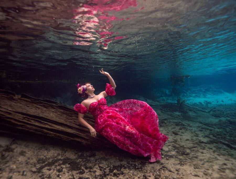 Woman poses at a submerged tree in a red-pink dress during an underwater modeling workshop